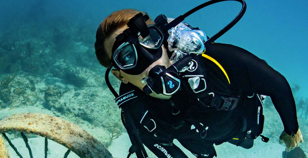 Plongeur en combinaison épaisse évoluant à moyenne profondeur dans une eau bleu-vert froide, respirant calmement avec son détendeur, faisceau de lumière naturelle filtrant depuis la surface